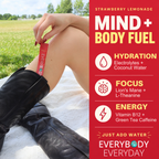A person outdoors on a white blanket wearing black boots and holding a red EveryBody Everyday energy stick packet. The right side features a red informational panel with text for 'Strawberry Lemonade' and product benefits: hydration, focus, and energy.